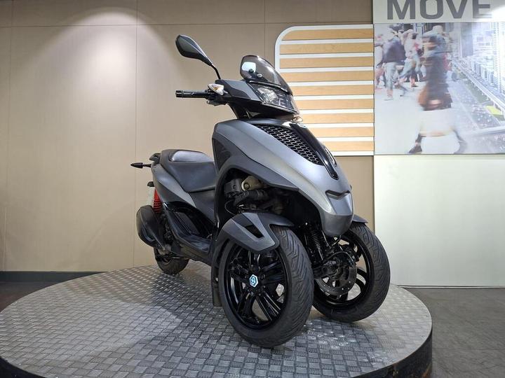 A sleek, gray three-wheeled scooter displayed on a circular platform in a modern showroom, with a textured metal floor and a backdrop featuring a busy urban scene.