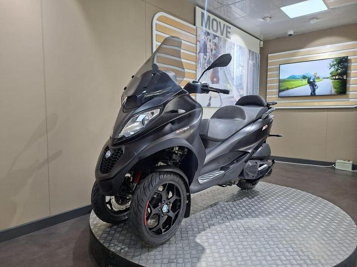 A black three-wheeled scooter displayed on a circular platform inside a modern showroom, with a wall featuring a "MOVE" sign and a monitor showing a scenic landscape in the background.