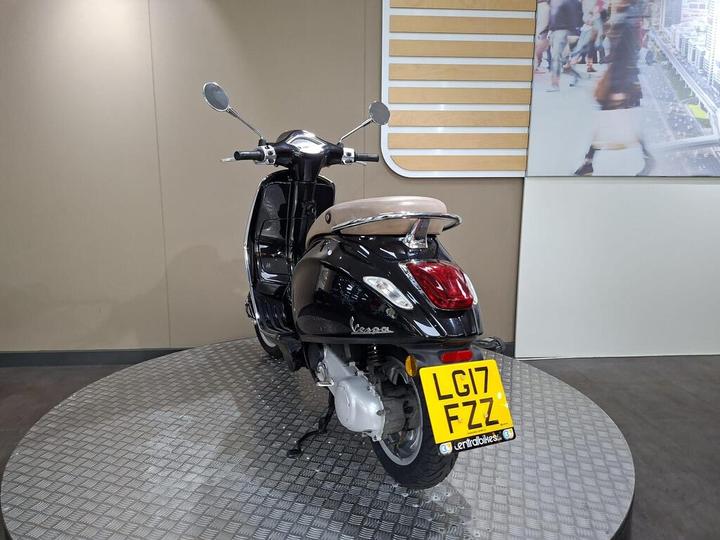 A black Vespa scooter with a beige seat, displayed on a circular platform, facing slightly to the right. The license plate reads "LG17 FZZ." A digital screen with a cityscape is visible in the background.