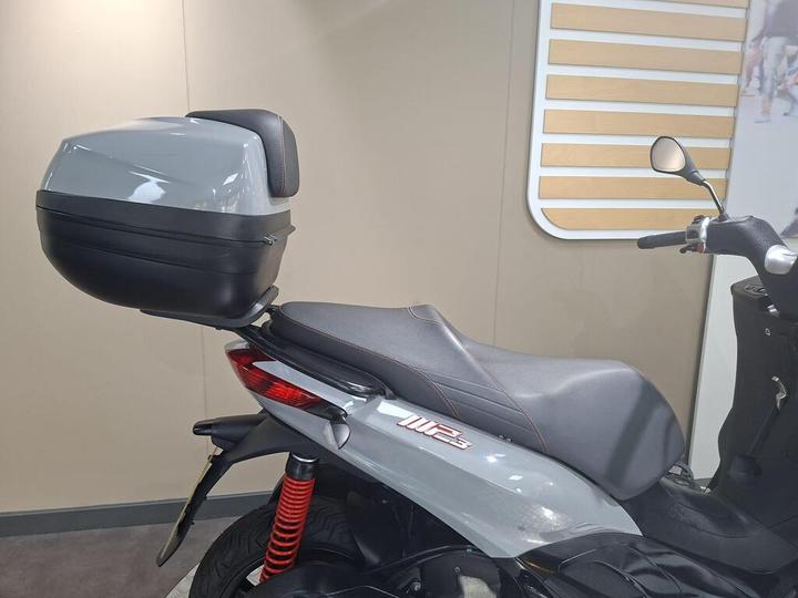 A grey scooter with a black top case, showing the seat and rear suspension, set against a neutral background.