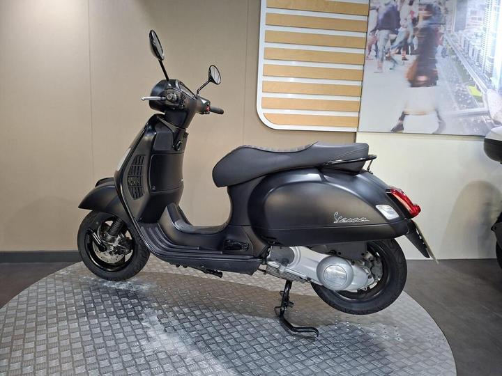 A matte black Vespa scooter is displayed on a circular metal platform, with a minimalist background featuring a blurred cityscape. The scooter is viewed from the side, showcasing its sleek design and detailing.