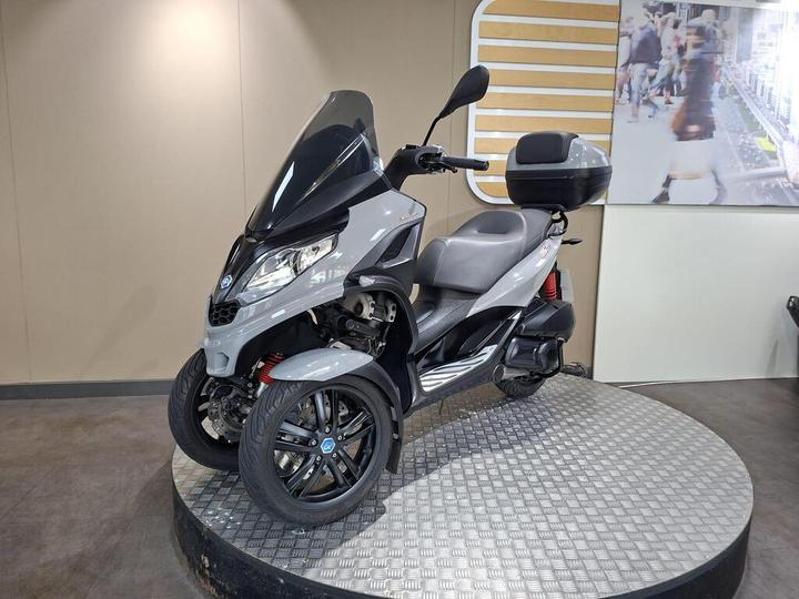A gray three-wheeled scooter parked on a circular platform, featuring a front shield and a storage compartment on the back, displayed against a neutral background.