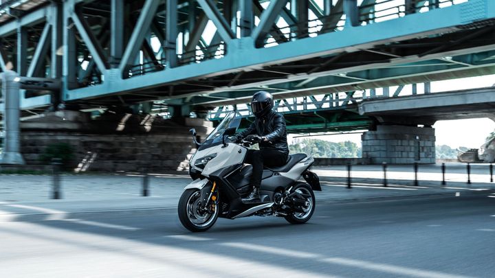 A motorcyclist in a black helmet and leather jacket rides a sleek, white scooter under a large bridge, with sunlight creating contrasting shadows on the pavement.