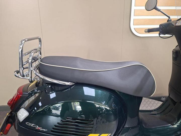 A close-up of a green scooter's rear section, featuring a sleek black seat with white piping and a chrome luggage rack attached behind it. The background is a plain light-colored wall.
