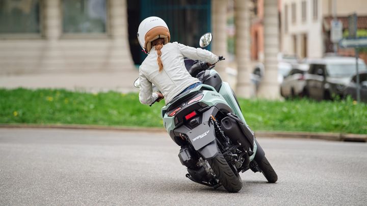 A rider in a white jacket and helmet is maneuvering a green scooter around a curve on a city street, with greenery and parked cars in the background.