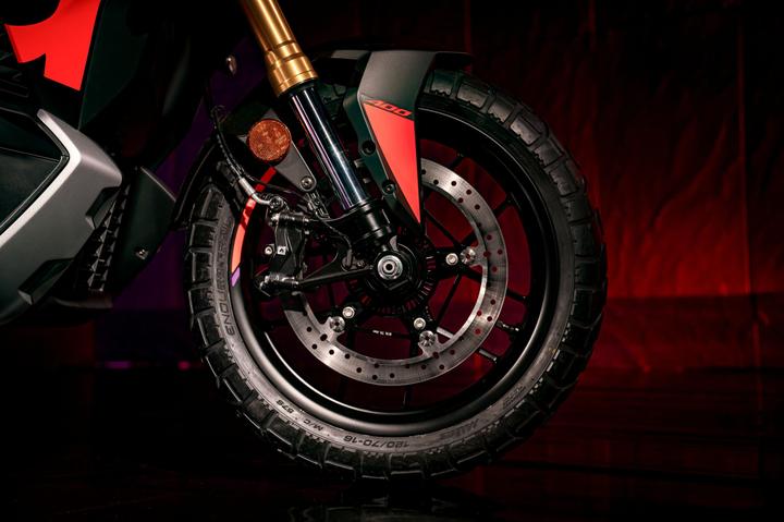 Close-up of a motorcycle's front wheel, featuring a detailed tread pattern, disc brake, and suspension elements, set against a gradient background with red and purple hues.