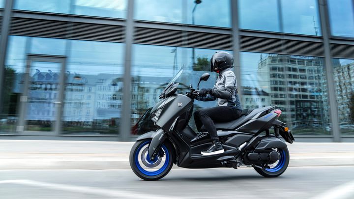 A rider in a helmet and a gray jacket is cruising on a sleek black Yamaha scooter with blue rims, passing by modern glass buildings reflecting the cityscape.