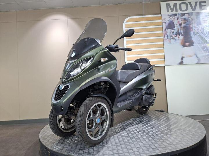 A green three-wheeled scooter displayed on a circular platform, with a transparent windscreen, in an indoor setting featuring a light-colored wall and a promotional sign in the background.