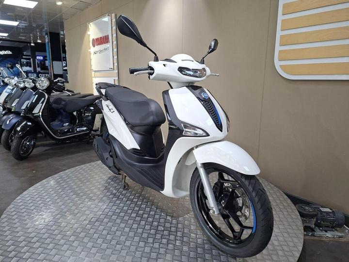 A white scooter with a sleek design is displayed on a diamond-patterned platform in a Yamaha showroom, surrounded by various motorcycles in the background.