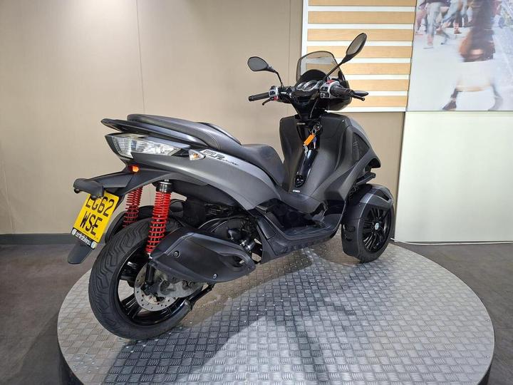 A black three-wheeled scooter displayed on a circular, textured platform with a neutral background. The rear view shows its distinctive design, red shock absorbers, and license plate reading "LG62 WSE."