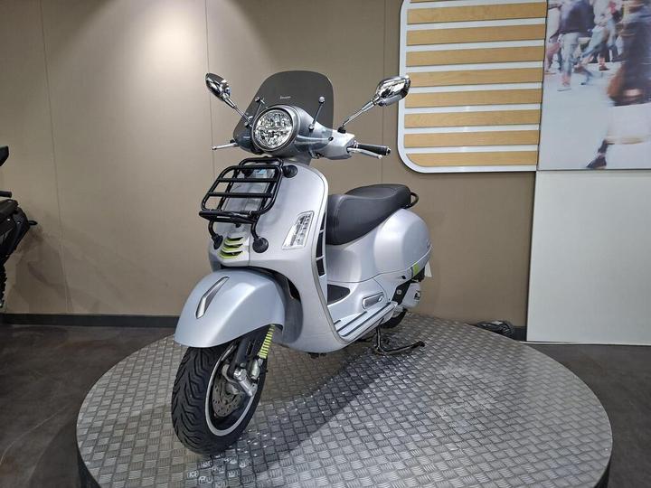 A silver scooter displayed on a circular metal platform in a showroom, featuring a front rack and chrome mirrors, with a light gray wall and a decorative panel in the background.