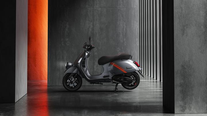 A sleek silver scooter is positioned at an angle against a backdrop of textured gray and orange walls, showcasing its modern design and streamlined form.