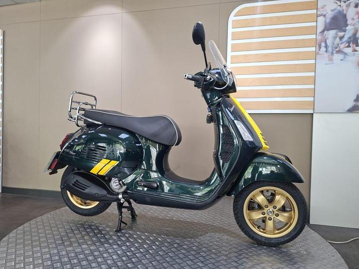 A green scooter with gold wheels and a yellow accent, displayed on a circular platform in a modern showroom setting.
