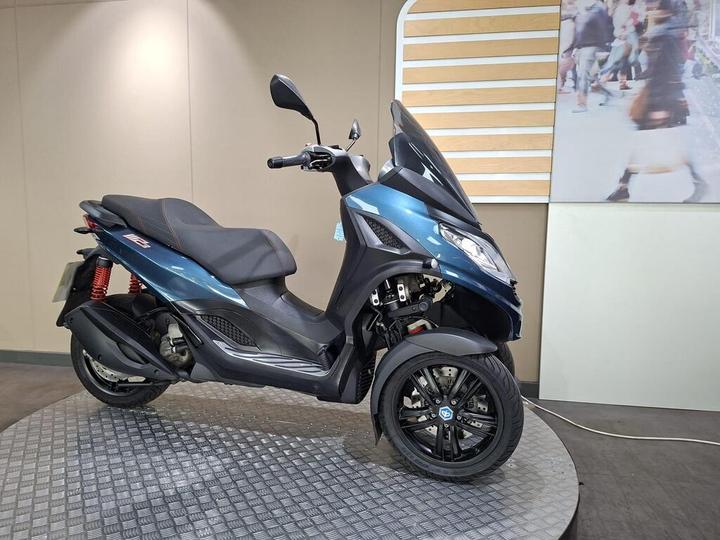 A blue three-wheeled scooter displayed in a showroom setting, featuring a sleek design, red rear suspension, and a circular textured platform beneath it.