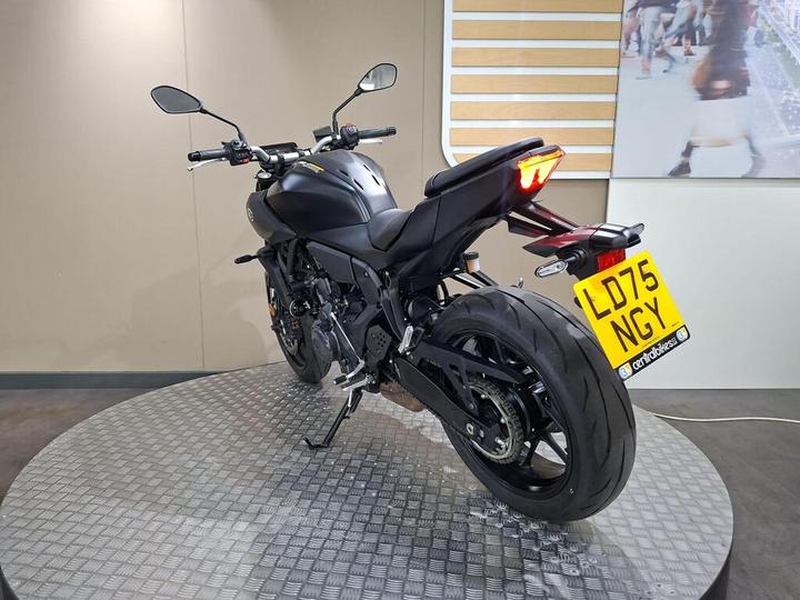 A black motorcycle is displayed from the rear on a circular platform with a textured surface. The bike has a sleek design, featuring mirrors, a distinctive tail light, and a license plate reading "LD75 NGY." The background is neutral, highlighting the motorcycle.