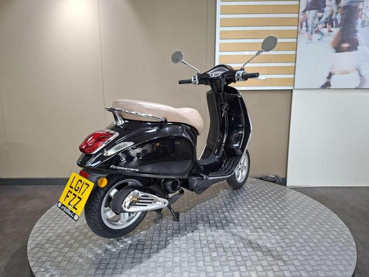 A black scooter with a brown seat is displayed on a circular metal platform, positioned against a neutral wall with horizontal wood paneling. The scooter features a rear view with clear mirrors and a visible license plate.