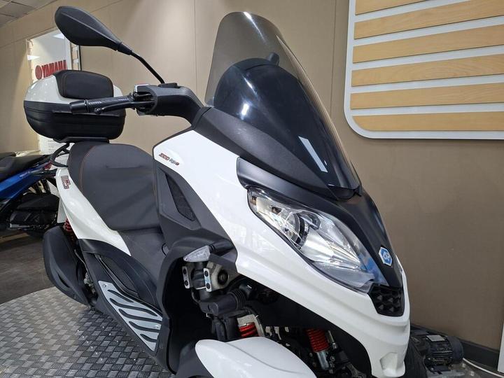 A close-up view of a modern white scooter with a streamlined design, featuring a windscreen, comfortable seat, and a red shock absorber. The background includes muted display elements of a store.