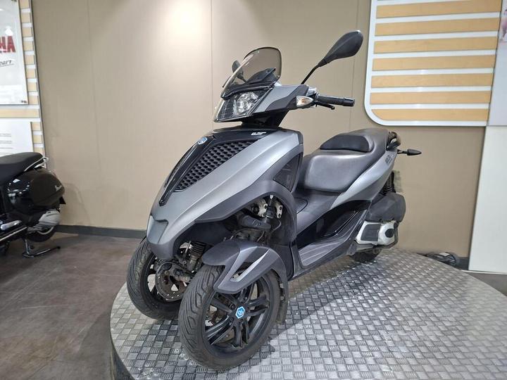 A grey three-wheeled scooter displayed on a circular metal platform in a showroom, with a plain beige wall in the background.