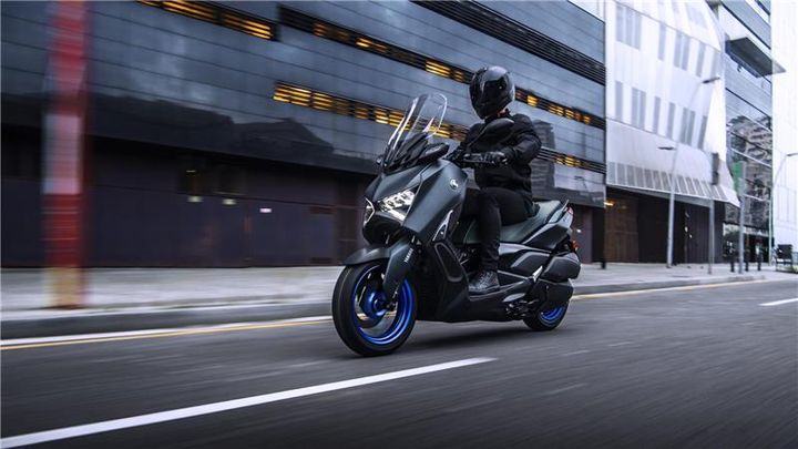 A rider in a black helmet and jacket navigates a sleek, dark motorcycle with blue accents on an urban street, with modern buildings in the background.