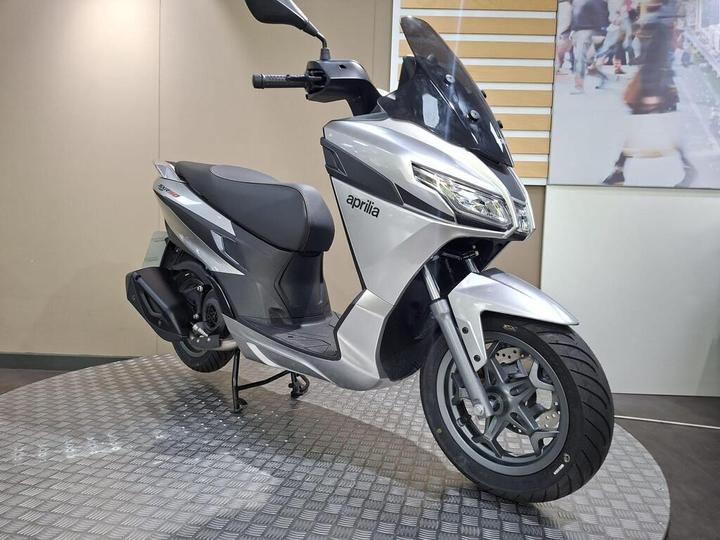 A silver Aprilia scooter displayed on a circular textured stand in a showroom setting. The scooter features a sleek design with a black seat and windscreen. A blurred background image of pedestrians is visible behind the scooter.
