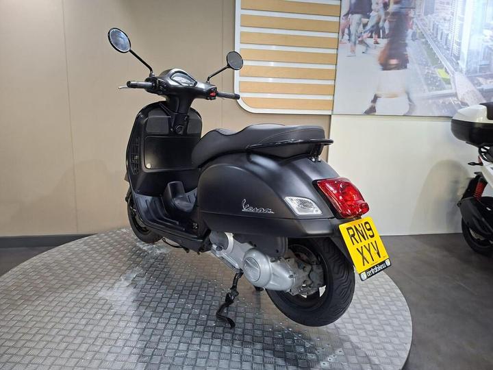 A black Vespa scooter parked on a circular metallic platform with its rear side visible, featuring a license plate displaying "RN19 XYV." The background shows a neutral wall and a partial view of another scooter.