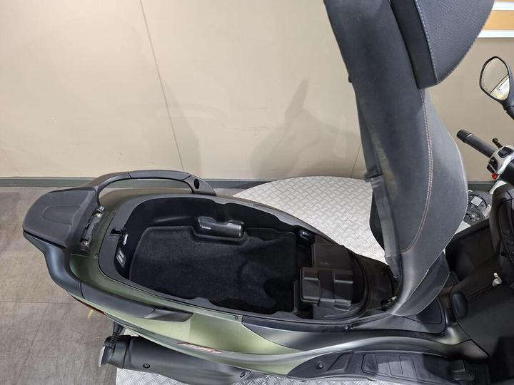 A top-down view of a scooter with the seat raised, revealing a storage compartment beneath. The interior is dark with a black carpet lining, and the scooter's body is a sleek green color.