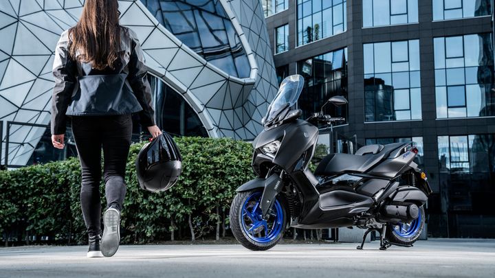 A person with long hair in a black and gray jacket walks away from the camera, holding a black motorcycle helmet, beside a sleek black scooter with blue wheels, set against a backdrop of modern glass buildings and geometric architecture.
