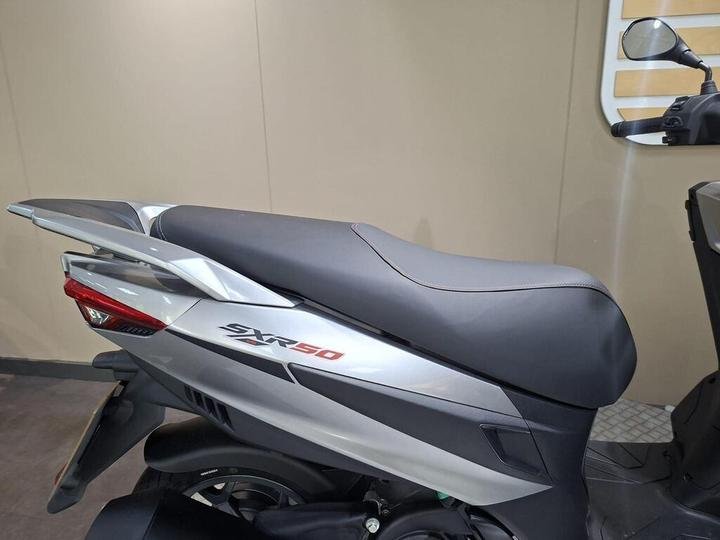 Close-up of the seat and rear section of a silver and black scooter, showcasing the "SXR50" model name and streamlined design elements against a neutral background.