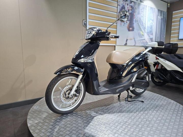 A black and beige scooter displayed on a rotating platform in a modern showroom, with a neutral background and another scooter partially visible on the right.