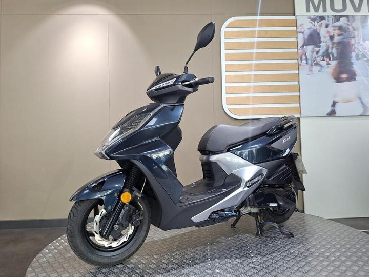 A dark-colored scooter is displayed on a textured platform, with a modern design featuring a sleek body and front headlight. The background is neutral, partially obscured by a promotional image of people walking.