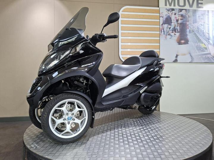 A black three-wheeled scooter displayed on a round platform with a textured surface, featuring a clear windscreen and a sleek design. The background includes a light-colored wall and a graphic with people walking.