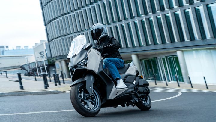 A rider in a black jacket and helmet is leaning into a turn on a modern scooter, set against a backdrop of contemporary glass buildings.