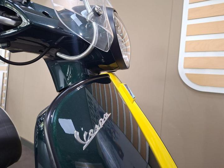 Close-up of a vintage Vespa scooter with a glossy green body and yellow accents, featuring a front headlight and clear windshield, set against a neutral background.