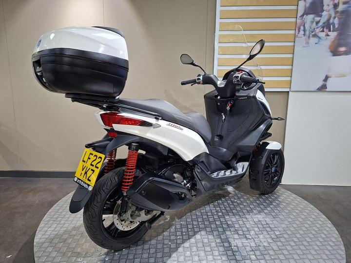 A three-wheeled scooter with a white and black design, featuring a rear storage box and red shock absorbers, displayed on a circular patterned platform in a showroom setting.