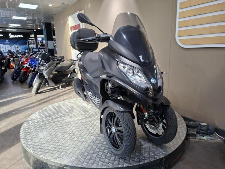 A black three-wheeled scooter displayed on a circular metallic platform in a motorcycle dealership, surrounded by other motorcycles.