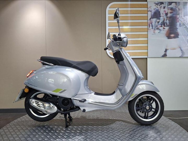Vespa Primavera 125 2025 A silver scooter is displayed in profile against a neutral backdrop with a graphic of a busy street scene in the background. The scooter features a sleek design with a black seat, chrome accents, and distinctive wheels.