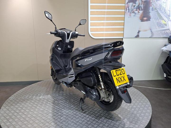 A sleek black scooter with a prominent rear end and a yellow license plate, displayed on a circular platform in a showroom with a neutral background.