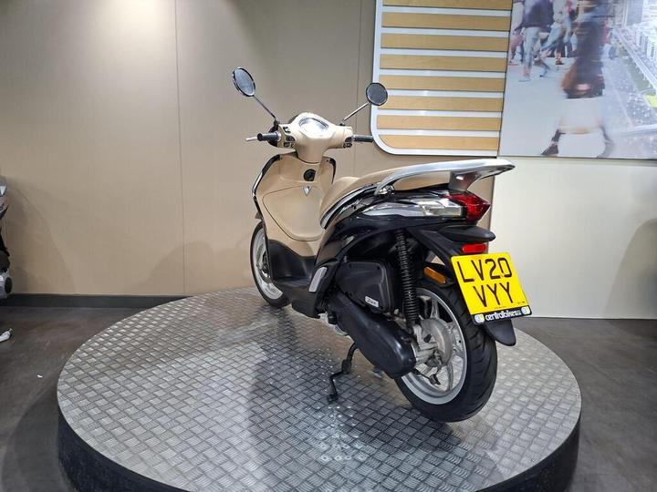 A beige scooter displayed on a circular pedestal in a showroom, with a visible license plate reading "LV20 VYY." The background features a minimalist wall and a large screen displaying city imagery.