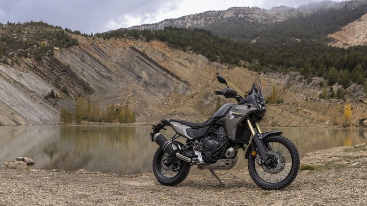 A gray adventure motorcycle stands on a rocky shore beside a calm lake, with layered mountains and dense forest in the background under a cloudy sky.