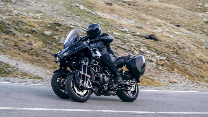 A motorcyclist in black gear rides a two-wheeled motorcycle with a large windshield and side panniers on a winding road surrounded by a rocky landscape.