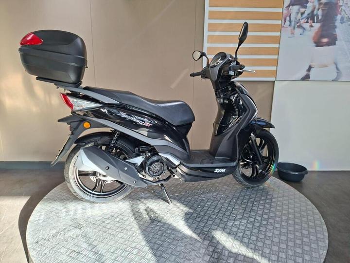 A black scooter displayed on a circular platform, with a rear top case attached, set against a light-colored backdrop.