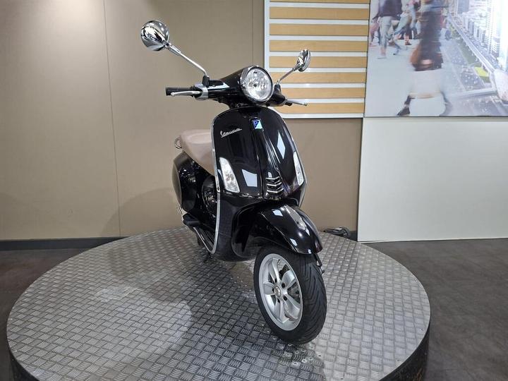 A black Vespa scooter with a beige seat, positioned on a circular display stand with a textured surface, against a plain background with minimal decor.