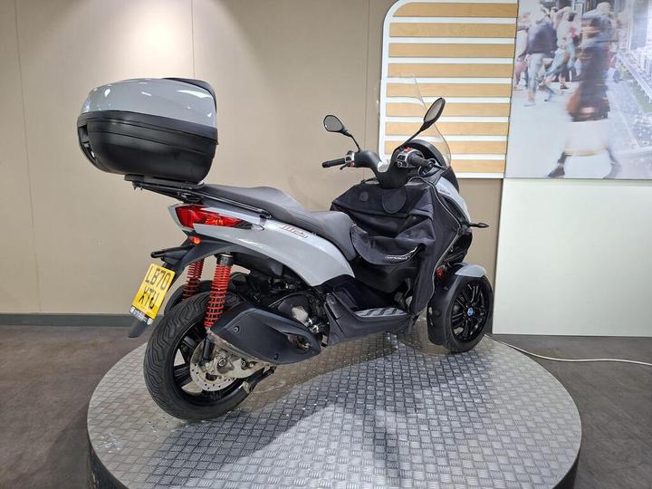 A three-wheeled scooter with a sleek silver and black design, featuring a rear storage trunk, positioned on a circular display with a beige wall in the background.