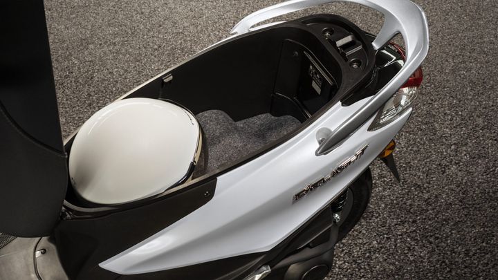 A top-down view of a scooter's storage compartment, featuring a white helmet inside, with a textured gray surface lining the bottom. The exterior is a sleek white and black design, highlighting the scooter's modern look.