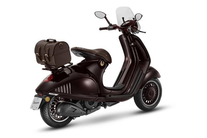 A dark brown Vespa scooter with a stylish design, featuring a brown leather bag attached to the rear, a clear windscreen, and distinctive wheel patterns, positioned on a white background.