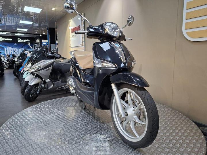 A black scooter with a beige seat is displayed on a circular metal platform in a showroom, surrounded by other motorcycles and scooters.