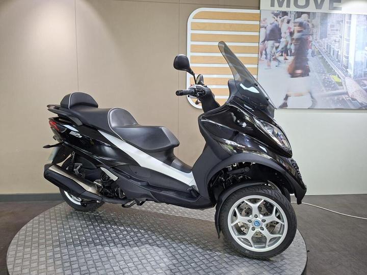 A black and silver three-wheeled scooter displayed on a circular platform, with a modern backdrop featuring an urban scene.