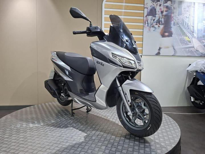 A silver Aprilia scooter displayed on a circular platform, featuring a tall windscreen and modern design elements, with a blurred backdrop of people and urban scenery.