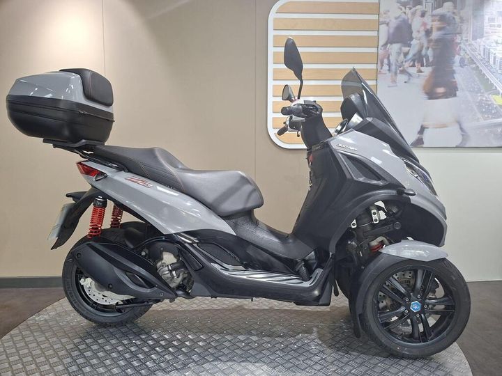Piaggio MP3 300 HPE Sport (2021) A gray three-wheeled scooter with a large top case, positioned on a textured metal surface in a modern showroom with a blurred background featuring people.