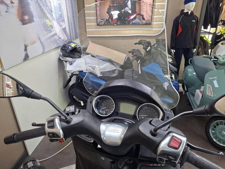 A view from the handlebars of a scooter, featuring a windshield, gauges, and controls, with various motorcycle gear and other bikes visible in the background.